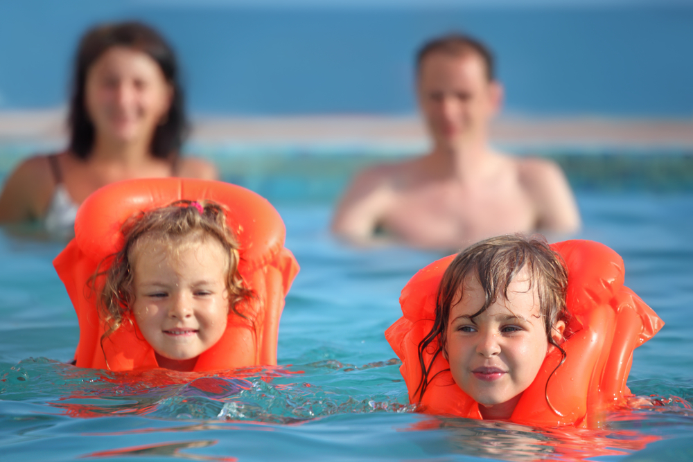 2 children swimming wearing life jackets with parents in background