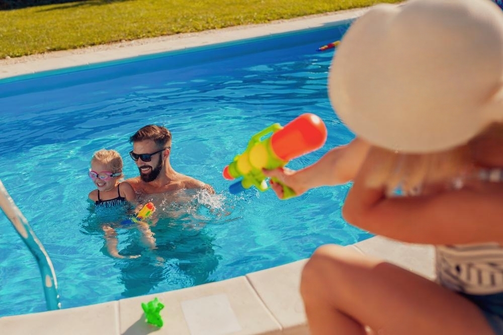 Parents and child playing with water squirt guns at a backyard swimming pool, enjoying quality family time in the sun.