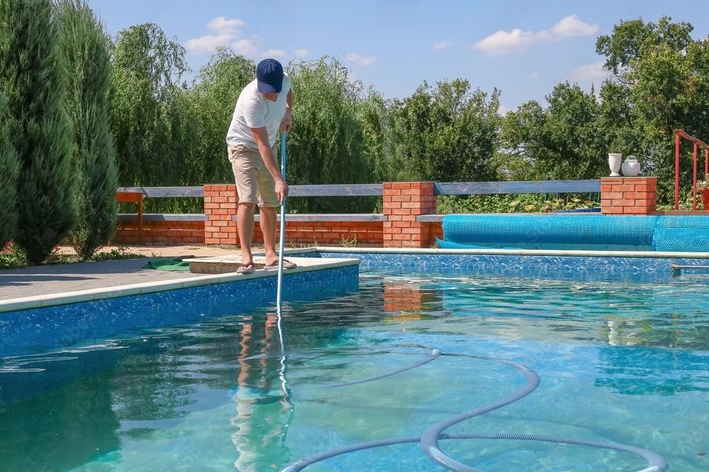 Man cleaning an outdoor swimming pool with a vacuum tube cleaner during the summer, preparing the pool for the season.