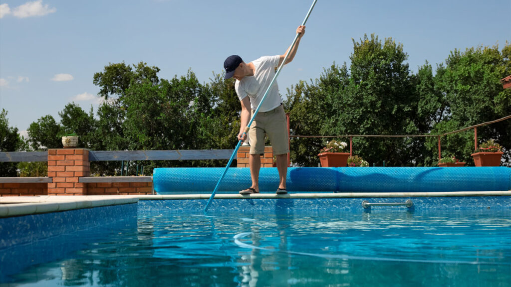 Man cleaning a sparkling outdoor swimming pool with a long vacuum pole, maintaining pool cleanliness on a bright sunny day.