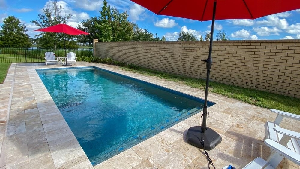 Relaxing backyard swimming pool with red umbrellas, bordered by a brick wall and light stone deck, featuring comfortable patio seating.