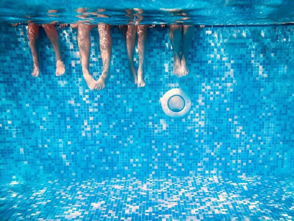 Children's and adults' legs underwater in a clear blue swimming pool with a tiled bottom.