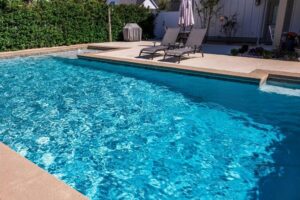 Rectangular backyard swimming pool with clear blue water and tan concrete edges, surrounded by a privacy hedge and lounge chairs.