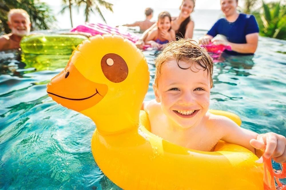 A smiling boy in a yellow inflatable duck float ring in a swimming pool with his family in the background, enjoying a sunny day together.