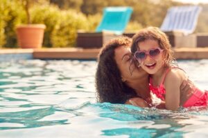 A mother laughing and holding her daughter in a swimming pool on a sunny day. The daughter is wearing heart-shaped sunglasses and a pink swimsuit, enjoying the fun moment.
