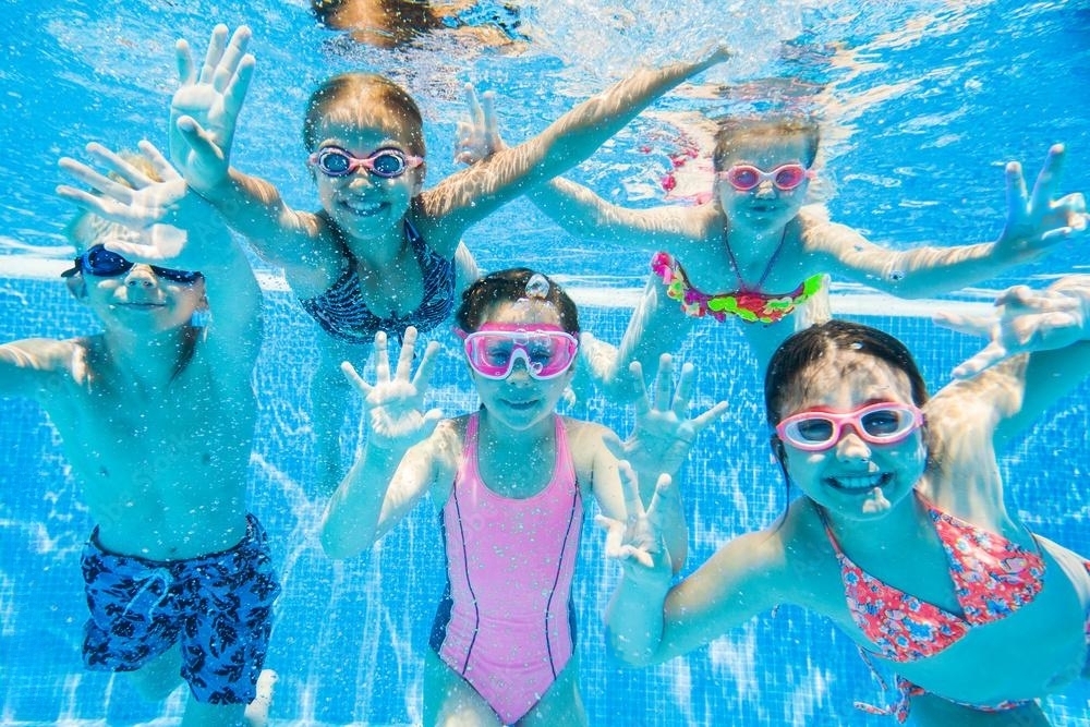 Five young children swimming underwater in a pool, wearing goggles and smiling as they wave at the camera, capturing a playful and energetic moment.