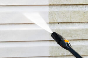 Close-up of a pressure washer cleaning dirt and grime off white vinyl siding, showing a clear before and after result.