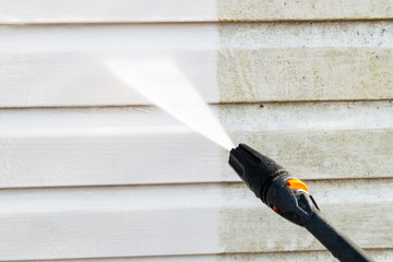 Close-up of a pressure washer cleaning dirt and grime off white vinyl siding, showing a clear before and after result.