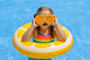 A young girl in a rainbow swimsuit floats in a clear blue swimming pool, smiling while holding orange halves over her eyes like goggles. She is inside a lemon-themed inflatable ring, representing summer fun and clean, inviting pool water thanks to swim n fun pool cleaning services