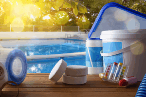Pool repair tools and chemicals on a wooden deck beside a clear blue pool, with sunlight filtering through nearby trees