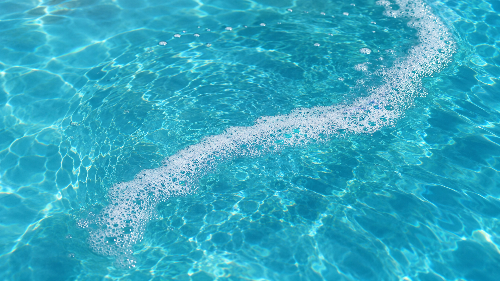 Foamy pool water Orlando shown by a delicate crescent of fine white bubbles floating across clear turquoise pool water in bright summer sunlight.